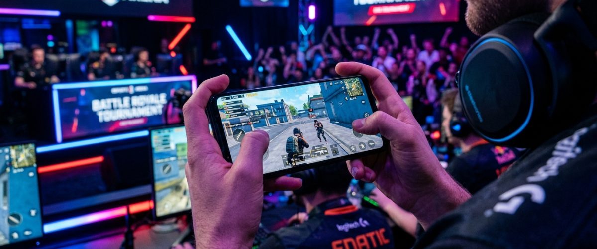 Hands holding smartphone showing battle royale mobile game with gaming setup