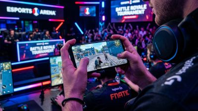 Hands holding smartphone showing battle royale mobile game with gaming setup
